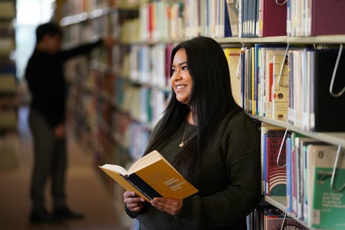 A female high school student leans on a library shelf while intently reading a book.