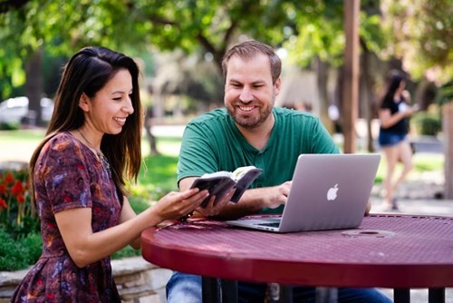 A man and woman sit at an outside table with a laptop and a bible